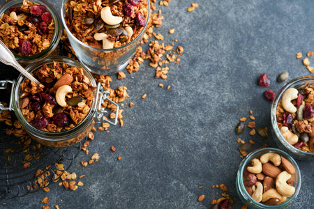 Homemade granola in glass jar with greek yogurt or milk and cashews, almonds, pumpkin with dried cranberry seeds in dark gray table background. Healthy energy breakfast or snack. top viewの写真素材