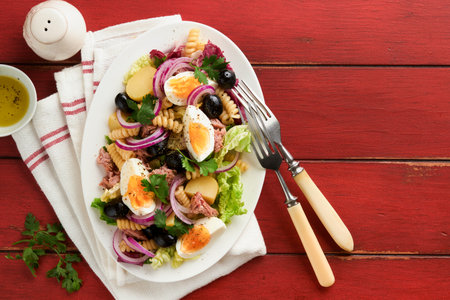 Tuna salad with pasta, eggs, potatoes, olives, red onions and sauce in white plate on old red rustic table background. Nicoise salad. french cuisine. top view. flat lay.の写真素材