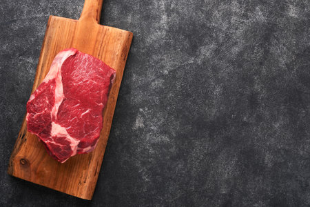 Raw beef steak. Fresh beef rib eye steak with fork rosemary, salt and pepper on piece of parchment paper on dark gray stone background. Shop advertising idea. top view. Mockup for design idea.の写真素材