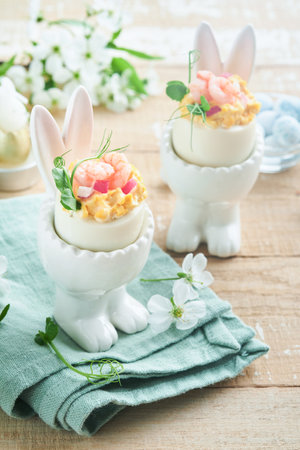 Stuffed or deviled eggs with yolk, shrimp, pea microgreens with paprika in rabbit-shaped stand for easter table decorate fresh cherry or apple blossoms on light background. Traditional dish for Easterの写真素材