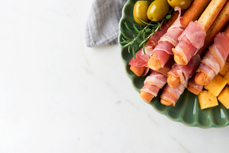 Slices of prosciutto or jamon. Delicious grissini sticks with prosciutto, cheese, rosemary, olives on green plate on dark background. Appetizers table with Italian antipasto snacks Top view copy spaceの写真素材