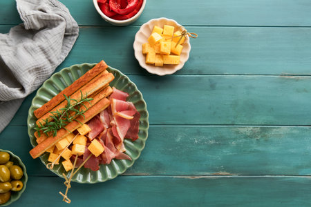 Slices of prosciutto or jamon. Antipasto Delicious grissini sticks with prosciutto, cheese, rosemary, olives on green plate on old wooden blue background. Appetizers table with Italian snacks Top viewの写真素材