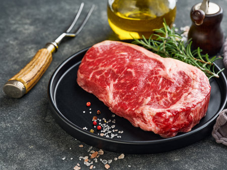 Raw beef steak on black plate with pepper, salt, rosemary and olive oil on black stone table with ample copy space. Dark background. Top view. Copy space.の写真素材