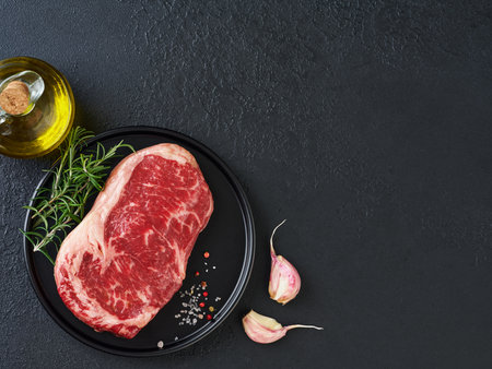 Raw beef steak on black plate with pepper, salt, rosemary and olive oil on black stone table with ample copy space. Dark background. Top view. Copy space.の写真素材