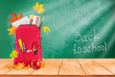 Red backpack for school with school supplies, red apple and flying autumn leaves on green blackboard background with copy space. Empty background. Back to school concept. Element for design.の写真素材