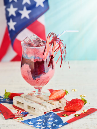 4th of July Holiday background. Punch with ice and strawberry pieces in glass and beautiful cocktail straw with fireworks against background of old turquoise board and sun rays.の写真素材