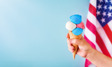 4th of July Holiday background. Mans hand holds colorful ice cream cone with red, white and blue scoops patriotic colors on blue background and American flag. Happy Independence Day concept. Mock up.の写真素材