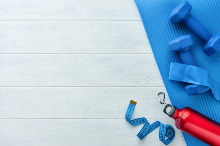Fitness or healthy lifestyle concept. Blue dumbbells, red bottle, exercise mat and measuring tape on white wooden background with copy space. Minimal sports accessories idea. Top view with copy spaceの写真素材