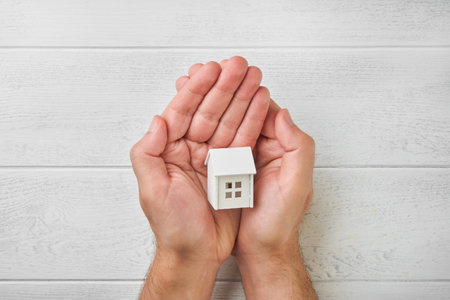 Male hands holding small toy house model on white wooden background. Family home, purchase and rent of apartments concept. Top view. Copy space.の写真素材
