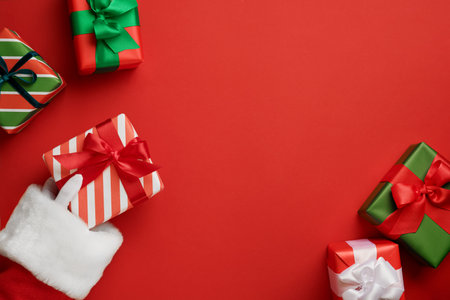 Santa Claus hand holding red and white striped gift box with red ribbon and other gifts on festive red background with copy space for text, Christmas card designの写真素材