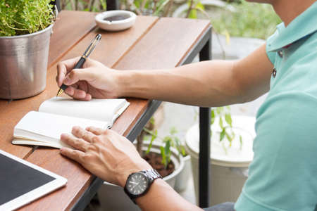 man writing on notebook with pen, on wooden table, outdoor sceneの写真素材