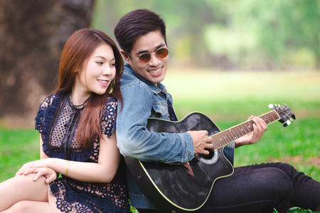 Asian couple in love playing acoustic song  guitar sitting on grass in the parkの写真素材