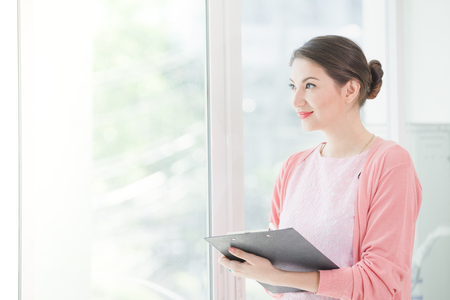 Business woman standing working near a window and holding clipboard at office.の写真素材