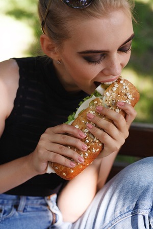 Hipster girt posing eating big sandwich. Outdoors lifestyle portrait