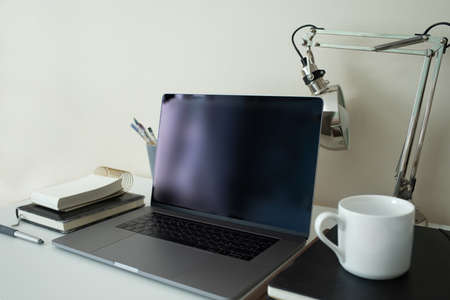 Modern stylish personal laptop with a black screen. Freelance artist or designer workplace. Clean display. Table lamp. Paint brushes. Notebooks for drawing and writing. White cup. Electronic gadgets.の写真素材