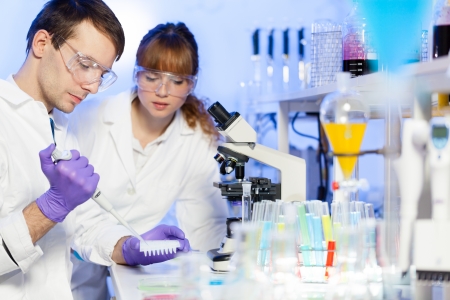 Young male researcher pipetting liquid in the 96 well microplate in the (forensics, microbiology, biochemistry, genetics, oncology...)laboratory. Female asistant science student watching and learning the protocol.の写真素材