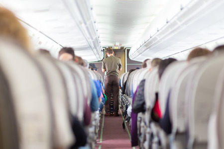 Interior of airplane with passengers on seats and steward walking the aisle.