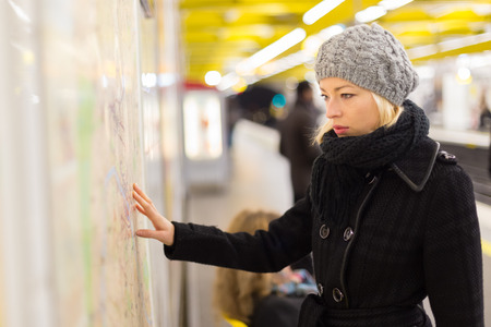 Casually dressed woman wearing winter coat, orientating herself with public transport map panel,...