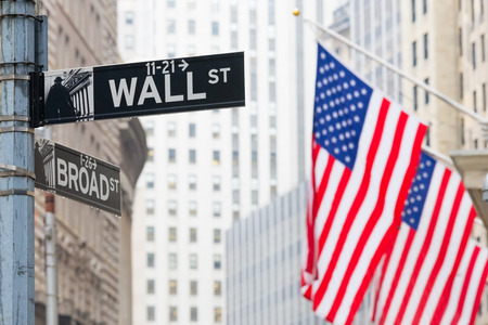 Wall street sign in New York with American flags and New York Stock Exchange background.