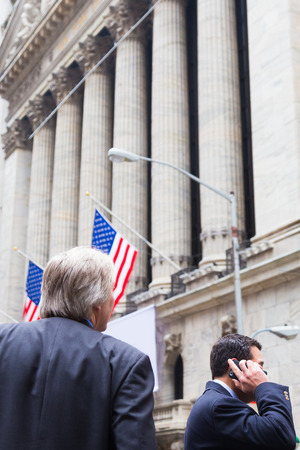 Businessman talking on the phone on Wall street in New York with American flags and New York Stock Exchange in background.の写真素材