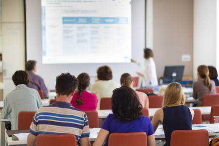 Speaker giving presentation in lecture hall at university. Participants listening to lecture and making notes.