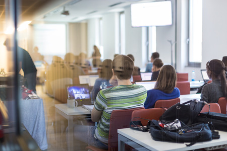 Workshop at university. Rear, trough the window, view of students sitting and listening in lecture hall doing practical tasks on their laptops.