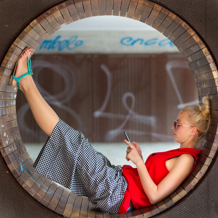 Casually dressed young cheerful lady wearing red sunglasses playing carelessly with her android smarthphone browsing trough social networks relaxing on  contemporary circular bench in city park.の写真素材