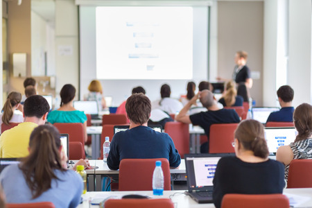 Workshop at university. Rear view of students sitting and listening in lecture hall doing practical tasks on their laptops. Copy space on white screen.