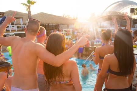 Sexy hot frinds dancing on a beach party event in sunset. Crowd dancing and partying at poolside in background. Summer electronic music festival. Hot summer party vibe.の写真素材