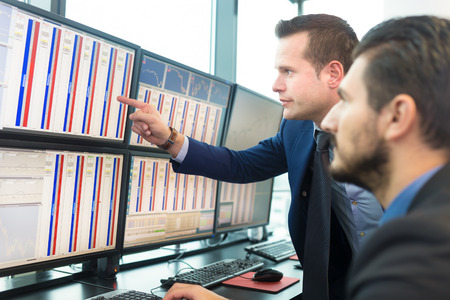 Businessmen trading stocks. Stock traders looking at graphs, indexes and numbers on multiple computer screens. Colleagues in discussion in traders office.