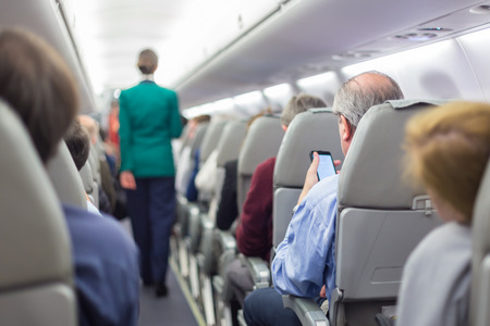 Interior of airplane with passengers on seats waiting to taik off. Stewardess in green uniform walking the aisle. Horizontal composition.の写真素材