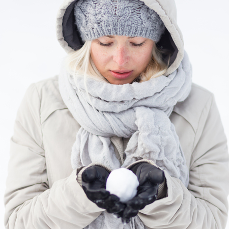 Cute casual young woman wearing glooves, woolen cap and scarf, holding icy snowball in cold winter time. Lady looking down at snowball.の写真素材