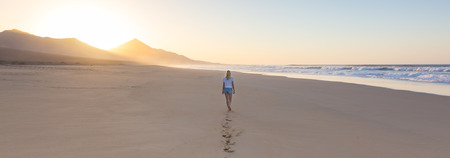 Woman walking on sandy beach in sunset leaving footprints in the sand. Beach, travel, concept. Copy space. Panoramic composition.の写真素材
