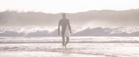 Silhouette of male surfer on the beach with the surfboard in sunset. Panoramic composition.の写真素材