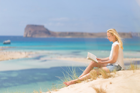 Relaxed woman enjoying sun, freedom and good book an beautiful sandy beach of Balos in Greece. Young lady reading, feeling free and relaxed. Vacations, freedom, happiness, enjoyment and well being.の写真素材