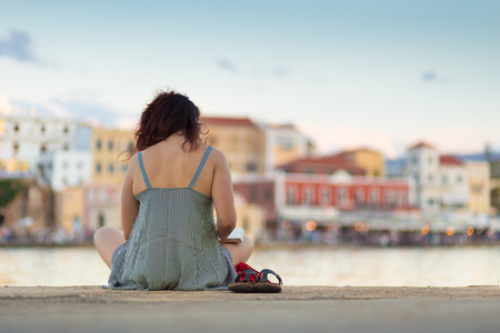 Rear view of woman sitting on a pier near sea port of Chania, Greece, reading book, enjoying...
