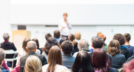 Business and entrepreneurship symposium. Female speaker giving a talk at business meeting. Audience in conference hall. Rear view of unrecognized participant in audience. Copy space on whitescreen.の写真素材