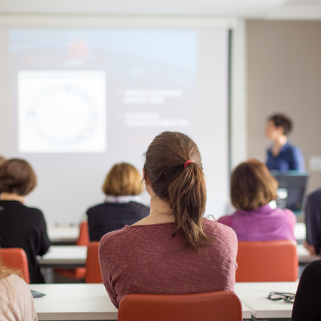 Female speaker giving presentation in lecture hall at university workshop. Rear view of unrecognized participants listening to lecture and making notes. Scientific conference event.の写真素材