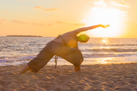 Silhouette of young woman practicing yoga on sandy beach at sunset.の写真素材