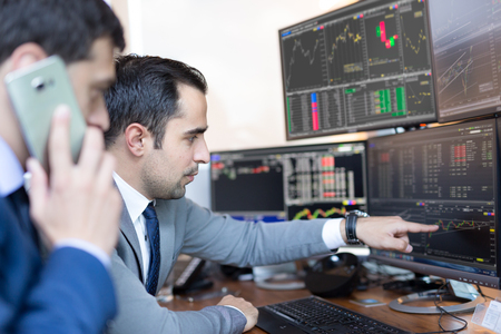 Businessmen trading stocks online. Stock brokers looking at graphs, indexes and numbers on multiple computer screens. Colleagues in discussion in traders office. Business success concept.の写真素材