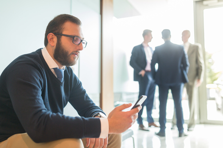 Businessman using smart phone while sitting and waiting in office building lobby. Business people talking in background.の写真素材