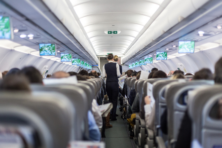 Interior of commercial airplane with flight attandant serving passengers on seats during flight. Stewardess in dark blue uniform walking the aisle. Horizontal composition.の写真素材