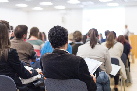 Business and entrepreneurship symposium. Female speaker giving a talk at business meeting. Audience in conference hall. Rear view of unrecognized participant in audience. Copy space on whitescreen.の写真素材
