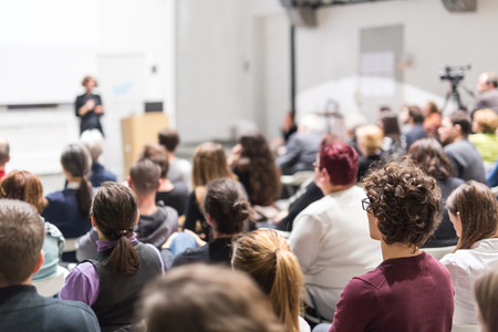 Female speaker giving presentation in lecture hall at university workshop. Audience in conference hall. Rear view of unrecognized participant in audience. Scientific conference event.の写真素材
