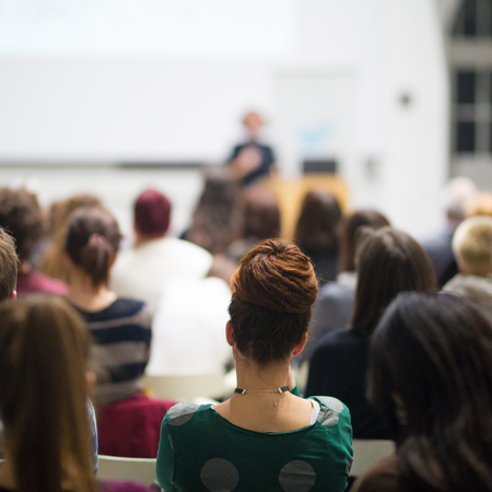 Female speaker giving presentation in lecture hall at university workshop. Audience in conference hall. Rear view of unrecognized participant in audience. Scientific conference event.の写真素材