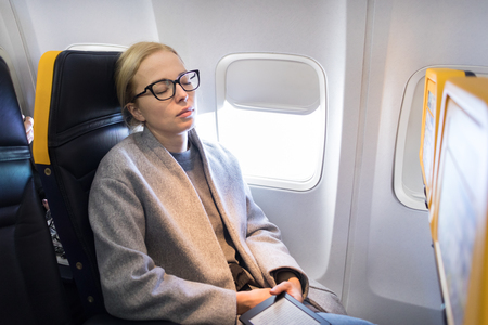 Tired blonde casual caucasian woman wearing glasses, holding digital e-reader, napping on seat while traveling by airplane. Commercial transportation by planes.の写真素材