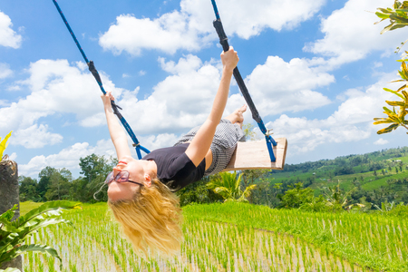 Happy caucasian female traveller swinging on wooden swing, enjoying summer vacation in pristine green nature at Jatiluwih rice terraces on Bali, Indonesia. Freedom and life enjoying concept.の写真素材