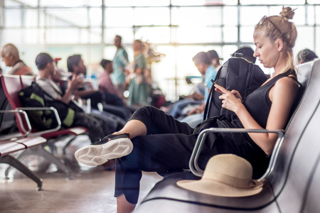 Casual sporty young blond female traveler using her cell phone while waiting to board a plane at the departure gates at the asian airport terminal.の写真素材