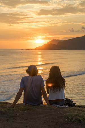 Young romantic couple on vacation sitting and watching colorful sunset over the sea coast.の写真素材
