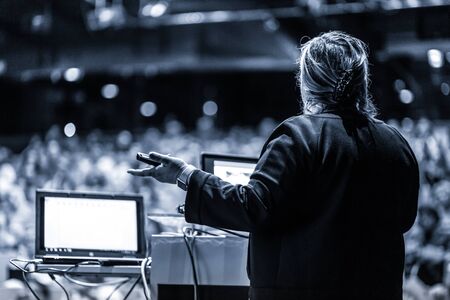 Female speaker giving a talk on corporate business conference. Unrecognizable people in audience at conference hall. Business and Entrepreneurship event. Black and white, blue toned image.の写真素材